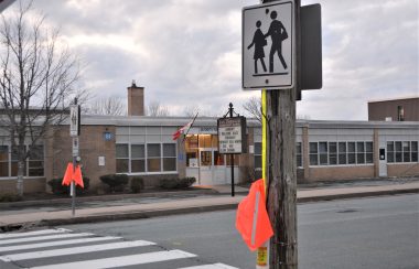 A crosswalk with orange flags hanging from post. Beyond the crosswalk is the entrance to a school.