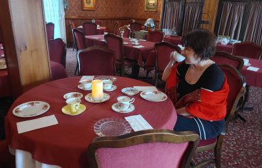 Coderre sitting at one of the tables in the dining room sipping on her tea. The tops of the tables have red table cloth/ You can see the tables set with tea cups and dishes.