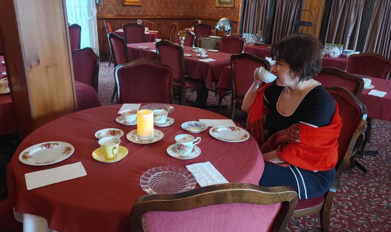 Coderre sitting at one of the tables in the dining room sipping on her tea. The tops of the tables have red table cloth/ You can see the tables set with tea cups and dishes.