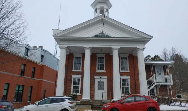 The old court house is made of brick and presents a typical European facade. The front of the building has five large windows. It has a large white roof over the entrance and is held up by four vertical pillars.