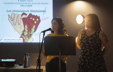 Pictured are one of the speakers standing behind the podium. She is accompanied by another woman and has a projected screen behind her.