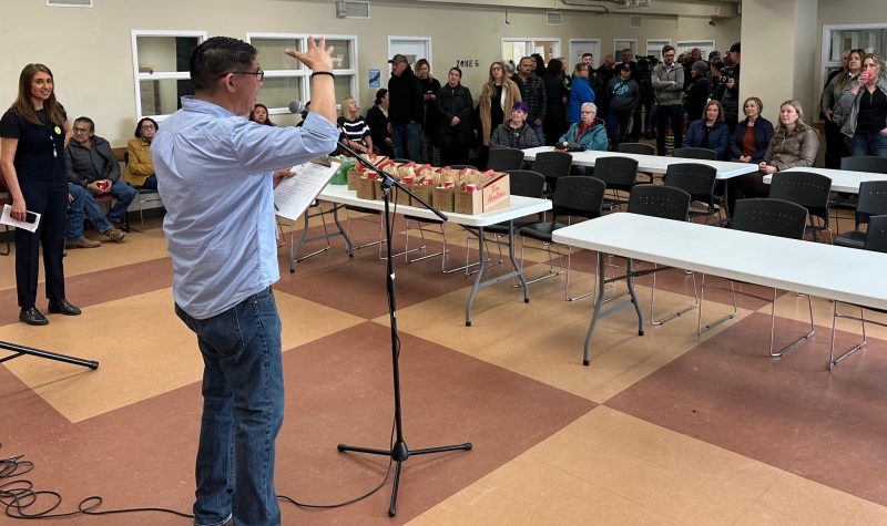 A speaker stands in front of a group of people, with a few fold-up tables in front of him. The photo is taken inside of the Lethbridge Wellness Centre.