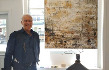 Michel Gamache standing beside a painting and what appears to be a sculpture of some sorts. A window serves as the background.