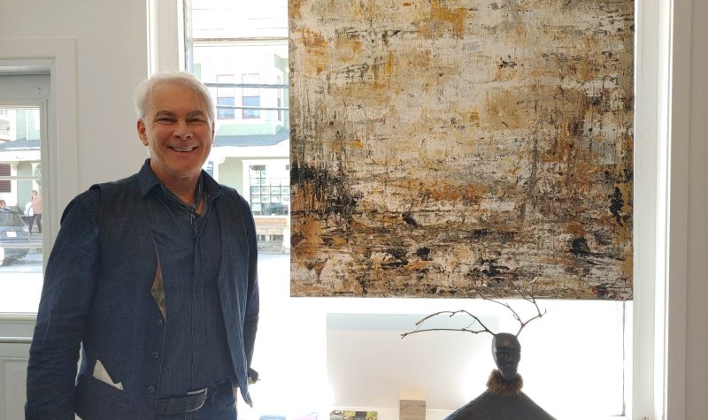 Michel Gamache standing beside a painting and what appears to be a sculpture of some sorts. A window serves as the background.