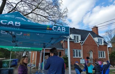 The grounds of CAB Cowansville. They are located in a brick building. In the photo you can see a blue tent written CAB Cowansville and some tables and chairs set up for their event. Some volunteers are also pictured sitting on the grounds.