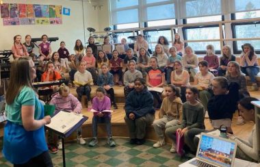 Pictured is Sherbrooke Elementary School's choir rehearsing with their teacher Tracey Rivette. The students are sitting on benches with Rivette standing in front of them to direct.