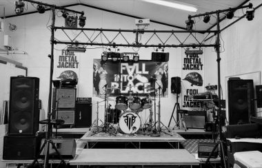 Une photo en noir et blanc d'une petite scène avec des haut-parleurs des deux côtés. Une batterie au centre avec d'autre équipement de musique. Derrière, il y a une affiche de « Fall Into Place ».