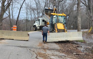 A municipal worker walks near a backhoe installing concrete barriers on a flooded section of road.