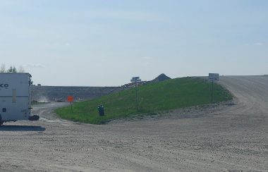 A truck is backing up on a gravel road next to a grassy berm at a landfill site.