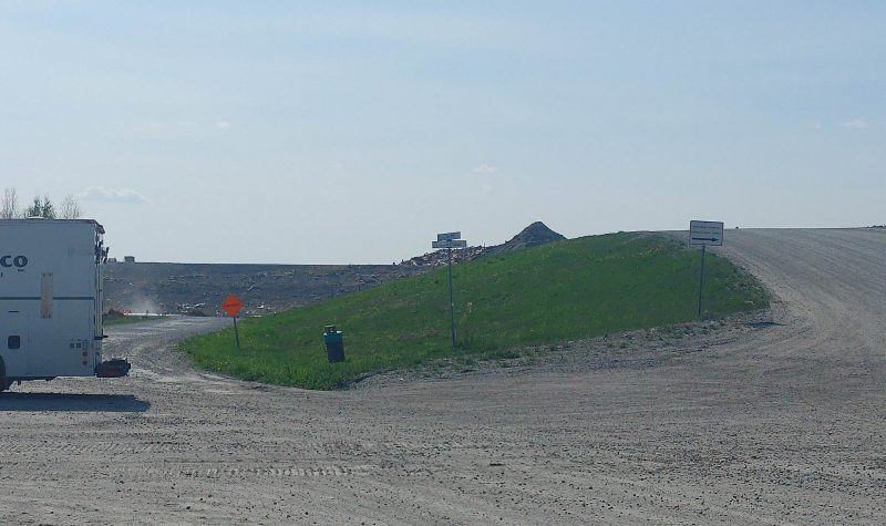 A truck is backing up on a gravel road next to a grassy berm at a landfill site.