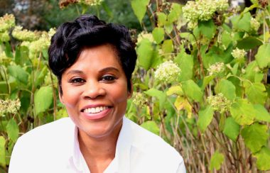 A woman smiling with green plants behind her.