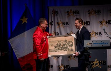Alexandre Cédric Doucet (extrème droit de la photo) donne une peinture à Mr. Terry Richardson, vêtu d'un manteau rouge. En arrière plan on aperçoit le drapeau acadien à gauche et le logo de la SANB sur fond blanc du milieu à la droite de la photo