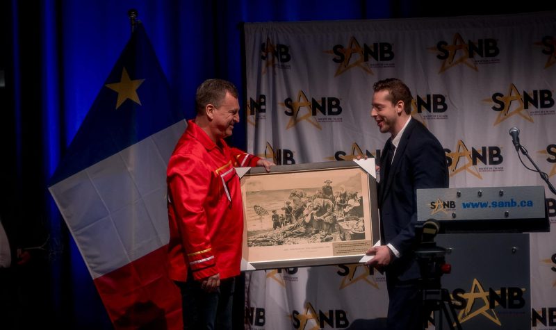 Alexandre Cédric Doucet (extrème droit de la photo) donne une peinture à Mr. Terry Richardson, vêtu d'un manteau rouge. En arrière plan on aperçoit le drapeau acadien à gauche et le logo de la SANB sur fond blanc du milieu à la droite de la photo