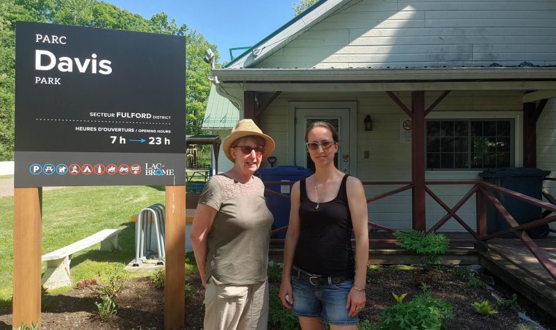 two people in summer clothing stand in front of a small white building and a sign that says Davis Park