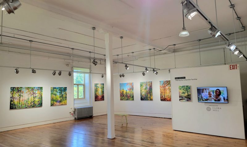 Art hangs in a well-lit white room with wooden flooring.