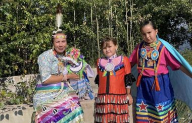Deux jeunes filles et une femme sont habillées en tenues traditionnelles autochtones. Elles sont très colorées. Elles sont dans la nature.