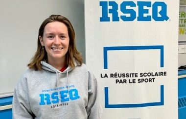 Une fille avec un chandail identifité RSEQ en bleu prenant la pose avec un sourire près d'une affiche identifitée RSEQ en lettre bleu