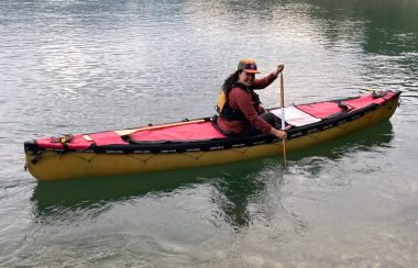 A woman wearing a red jacket and hat is sitting in a yellow canoe with red cover. Her paddle is in the water on the right hand side of the canoe.