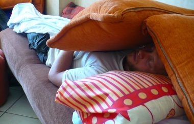 Man sleeping on a couch. Two pillows shield his face from the lights.