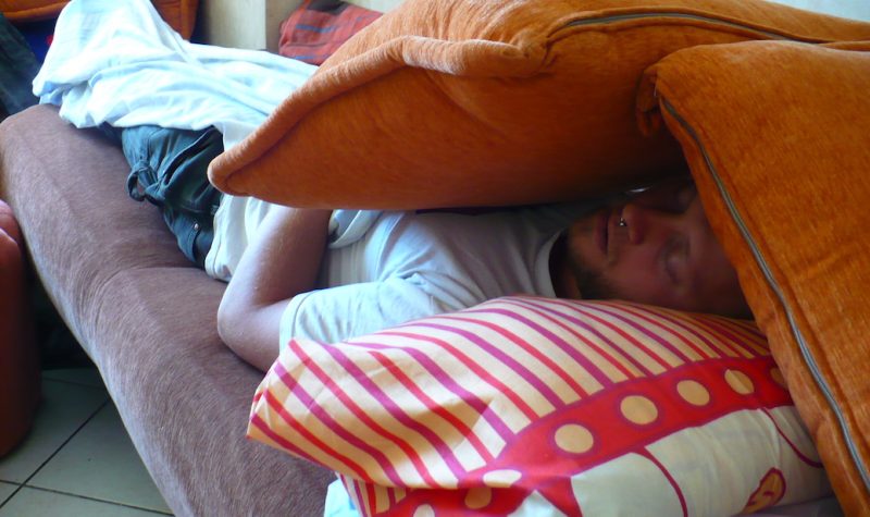 Man sleeping on a couch. Two pillows shield his face from the lights.