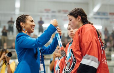 A woman in a blue jacket puts medal over young woman's head who is dressed in hockey equipment and an orange jersey.