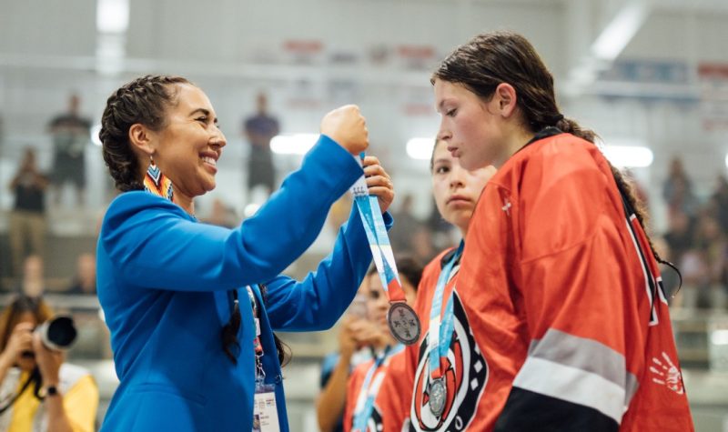 A woman in a blue jacket puts medal over young woman's head who is dressed in hockey equipment and an orange jersey.