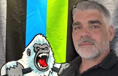 Dave Gagné, aux cheveux et à la barbe blanche prenant la pose devant une affiche des Nord-Côtiers