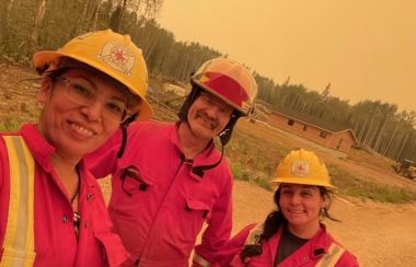 Trois personnes prennent un selfie. Ils sont habillés de combinaisons rouge et portent des casque jaunes de pompier. Ils sont à l'extérieur devant une forêt.