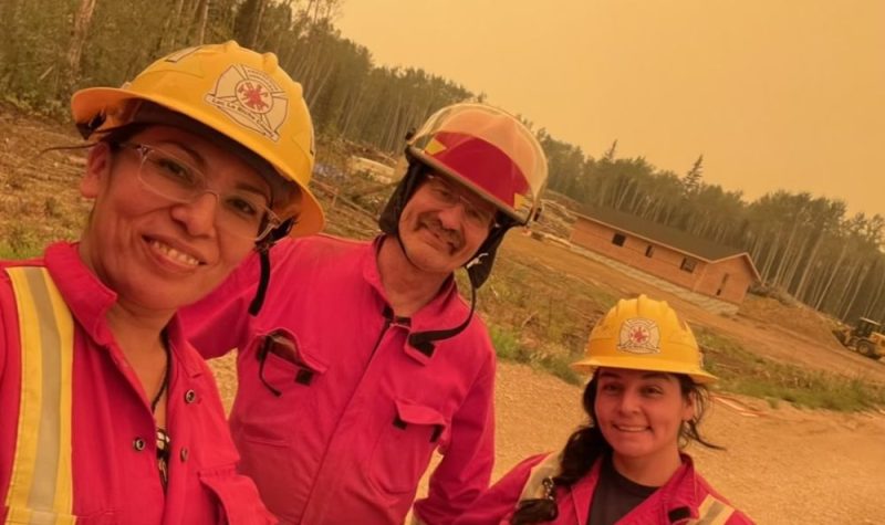 Trois personnes prennent un selfie. Ils sont habillés de combinaisons rouge et portent des casque jaunes de pompier. Ils sont à l'extérieur devant une forêt.