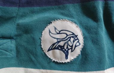 MVHS Vikings logo, outlined in dark navy blue and features a side-picture of a Viking with long hair and a helmet with horns. It is pictured on a textile background.