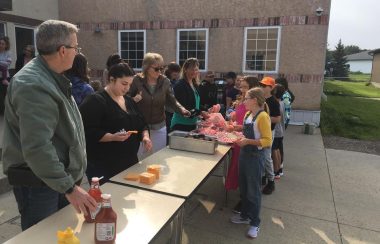 Une dizaine de personnes sont rassemblées autour de trois longues tables installées à l'extérieur. Sur les tables se trouve du fromage, des condiments et de la viande.