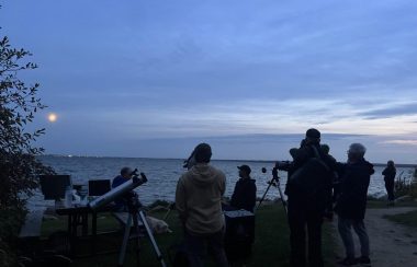 5 hommes sont debout devant un lac entourés de 3 télescopes. Un 6e homme leur fait face.