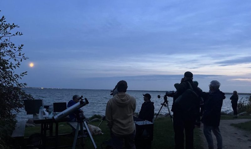 5 hommes sont debout devant un lac entourés de 3 télescopes. Un 6e homme leur fait face.