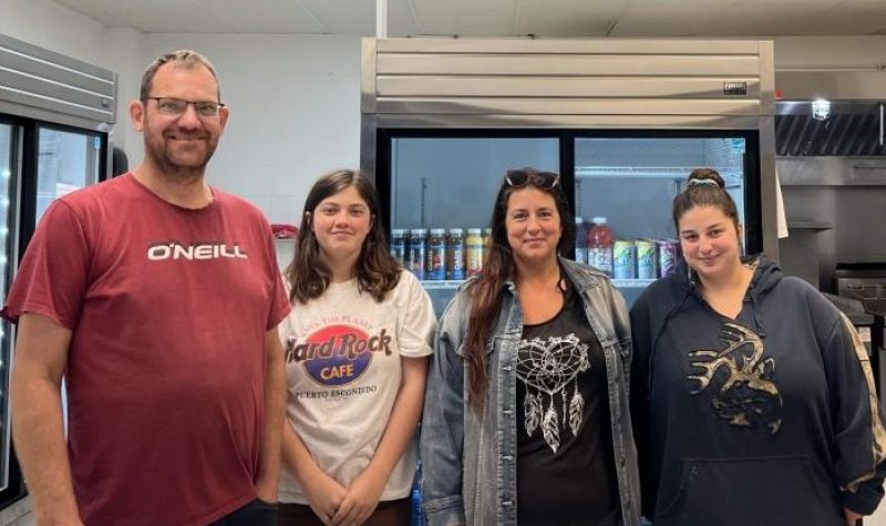 Un homme et trois femmes sourient à la caméra. Ils sont installés derrière un comptoir et devant un grand réfrigérateur.