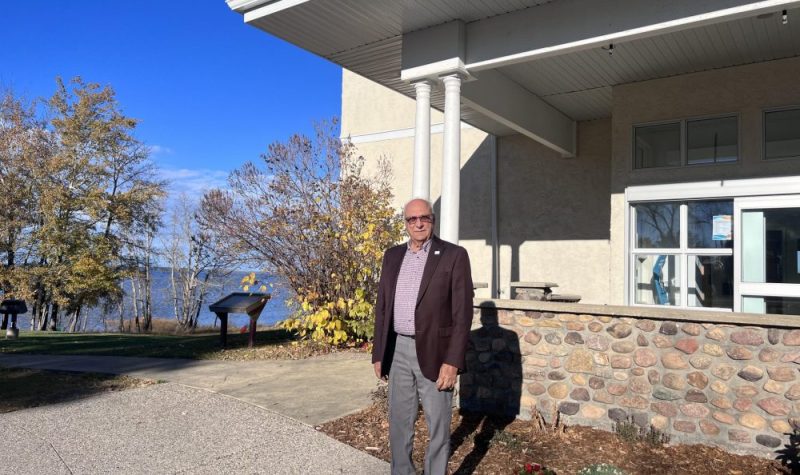 Un homme se tient devant un bâtiment beige. Au côté du bâtiment se trouve un lac et des arbres.