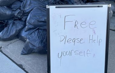 Une pancarte indique « Free Please Help Yourself ». La pancarte est située devant une montagne de sac de poubelles noirs.