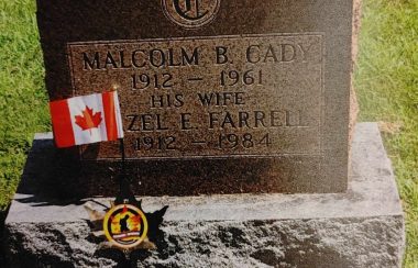 A medium sized brown granite grave stone with a small black marker with red black and yellow emblem in the centre. There is a Canadian flag next to the marker and gravestone.