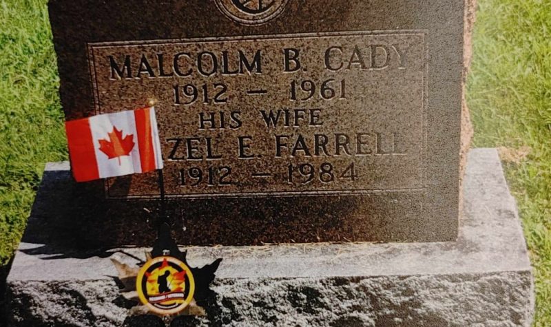 A medium sized brown granite grave stone with a small black marker with red black and yellow emblem in the centre. There is a Canadian flag next to the marker and gravestone.