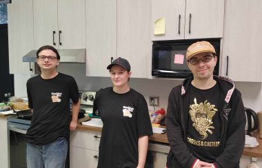 Three people wearing black tshirts are standing in front of white kitchen cupboards, counter a microwave oven and a stainless steel stove.
