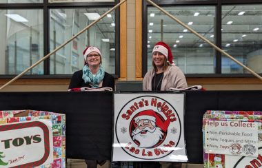 Deux femmes portent des chapeaux de Père Noël. Elles sont assises à une table décorée d'une affiche « Santa's Helpers »