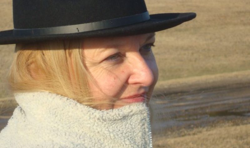 Une femme aux cheveux blond portant un chapeau noir et un manteau noir.