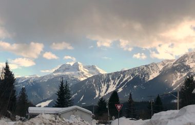 A snowy mountain scape. There is a snow covered building off to the left and large white clouds above.