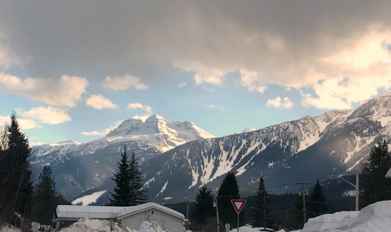 A snowy mountain scape. There is a snow covered building off to the left and large white clouds above.