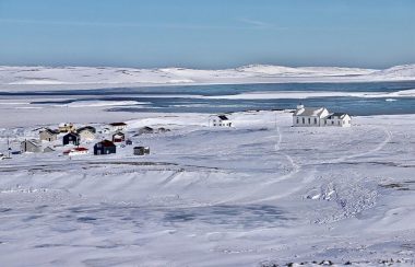 Un vaste paysage côtier enneigé, voire glacé, aux côtés d'une imposante baie adjacente au golfe du Saint-Laurent. Au loin, une église et quelques maisons regroupées.