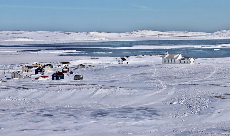 Un vaste paysage côtier enneigé, voire glacé, aux côtés d'une imposante baie adjacente au golfe du Saint-Laurent. Au loin, une église et quelques maisons regroupées.
