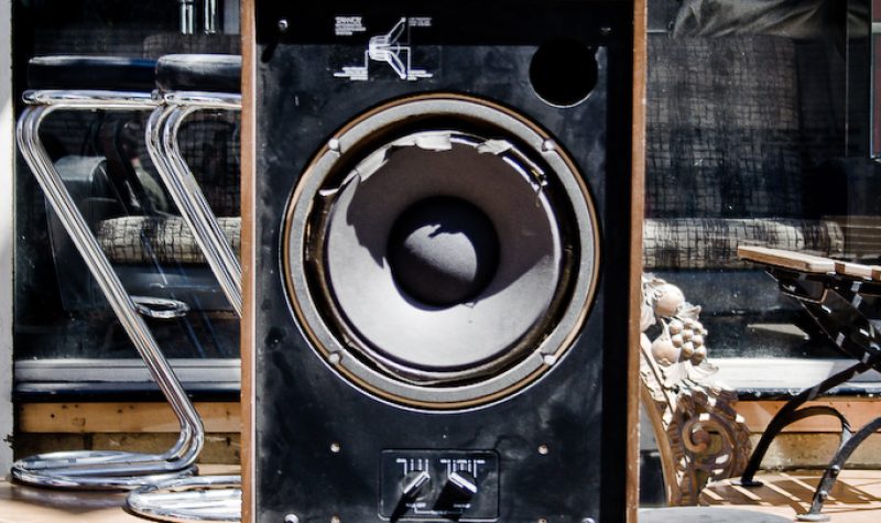 A large rectangular speaker with its front mesh removed, sitting on a baroque pillar amidst more chairs and furniture in the background.