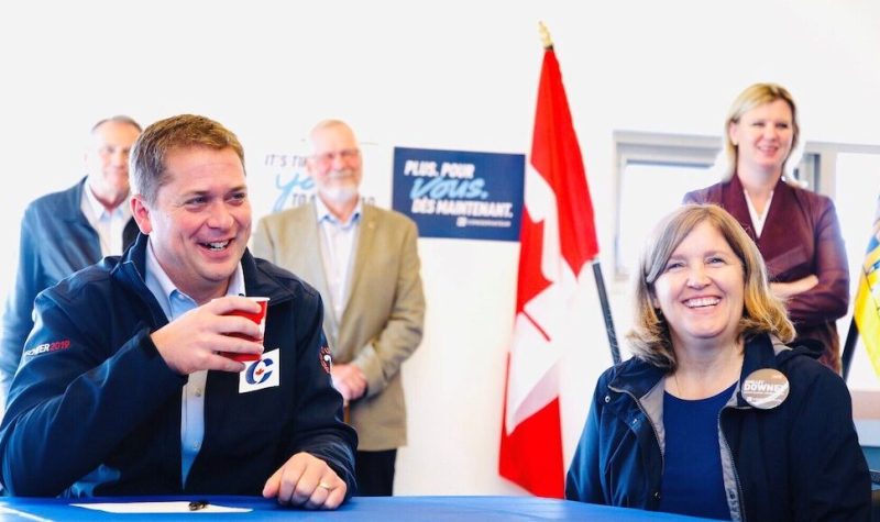 Andrew Scheer sits next to Shelley Downey at a meeting during the 2019 election season. There is a row of people behind them and a Canadian and British Columbian flag.