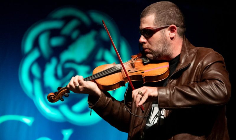 Un violoneux portant un gilet brun et des lunettes à soleil.