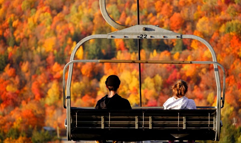Two people sitting on a chairlift making their way up the mountain with a view of colourful leaves.