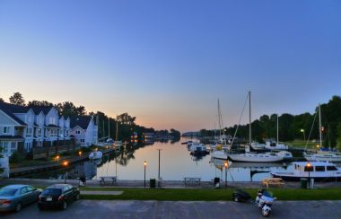 A waterfront at dusk, featuring sailboats and boats on the right side, and houses on the left side.